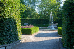 A sandy path cuts through hedges towards the circular inside of the garden, with a central fountain pond